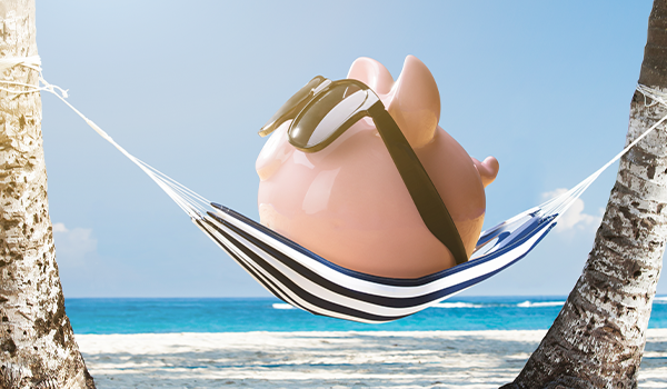 a pink piggy bank wears brown sunglasses and sits on a blue and white striped hammock which is hung between two trees. A sunny beach is in the background.